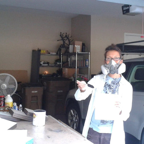 The artist in her garage studio, wearing a 3M mask and a lab coat while staining the sides of wood panels.