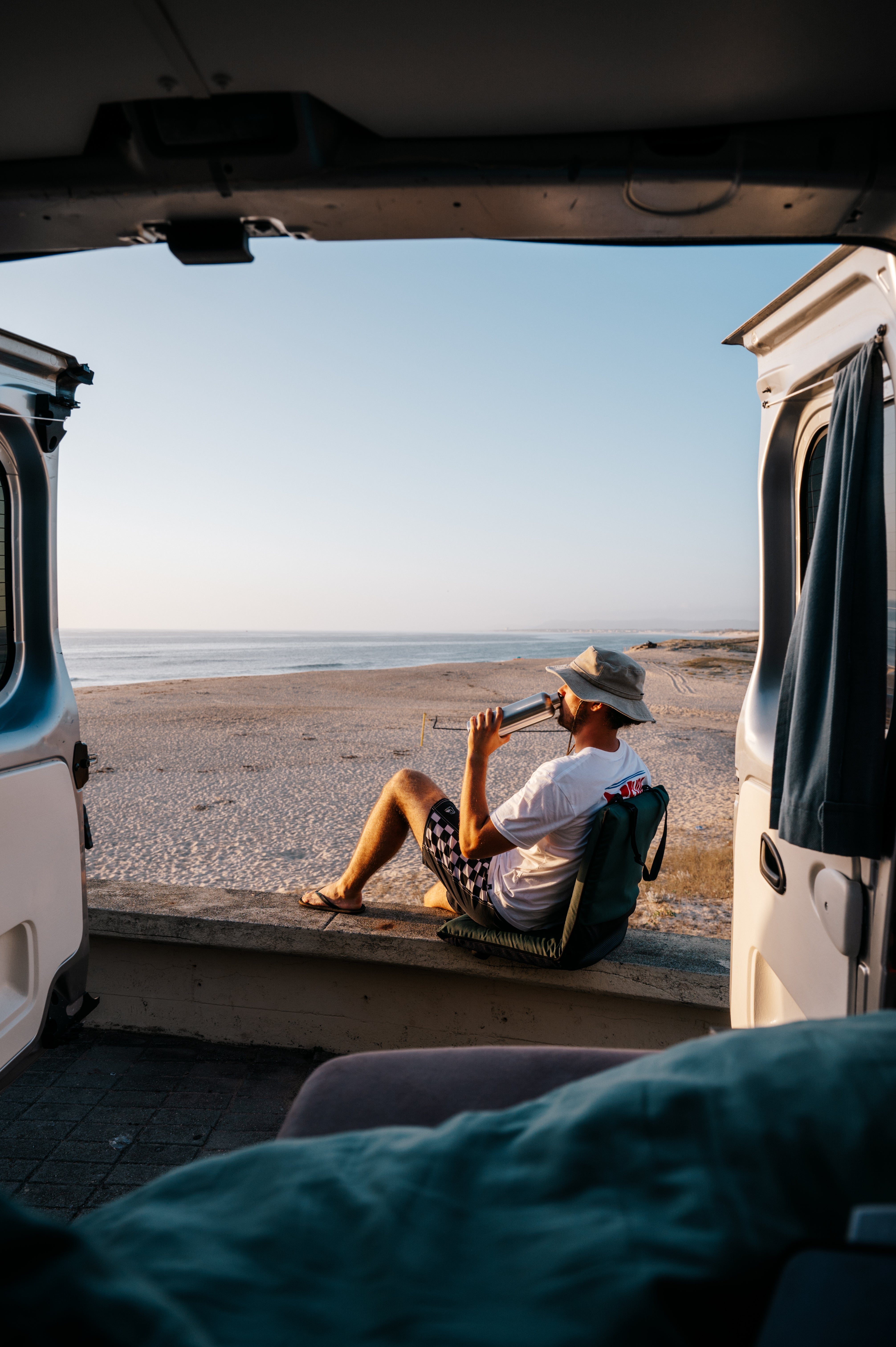 A guy drinking out of the Blockhuette stainless steel water bottle, photographed from the inside of a van, sitting at the beach