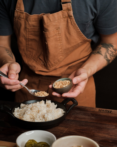 A mid-body shot of a man with tattooed arms clad in a brown apron, adding some garnishing to a pan of cooked rice