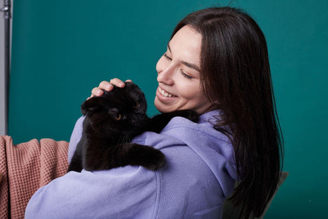 A medium close-up shot of a woman in a purple sweater cuddling with her black Scottish Fold in a green room