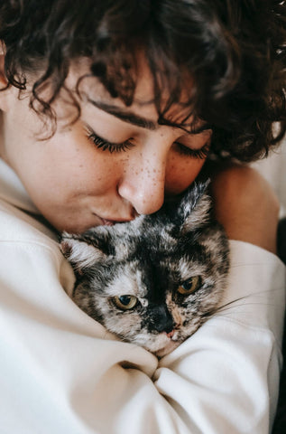A close-up of a woman with freckles kissing the head of a tortoiseshell wrapped in her arms
