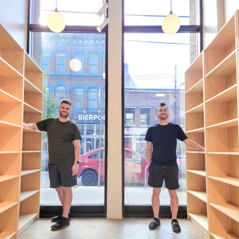 Tyler and Allan are standing in front of tall street view windows in their new storefront. It is empty still, except the new natural wood shelves they have installed.