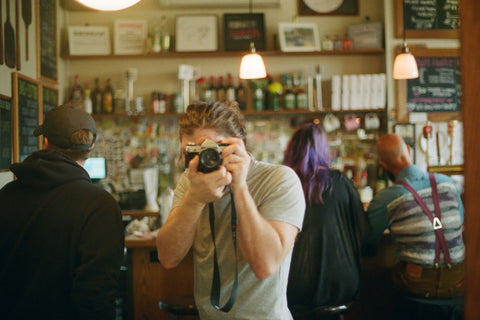 Caleb standing in a bar looking through his camera facing the camera