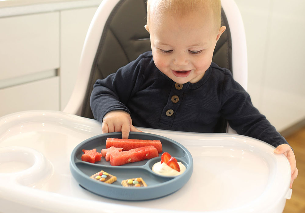 bébé garçon assis sur une chaise haute mangeant des fruits provenant d'une plaque d'aspiration en silicone avec séparateurs