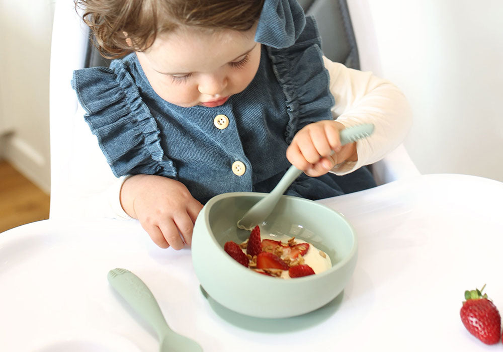 toddler girl in jeans sitting in a high chair and eating yogurt and strawberries with a silicone spoon from the suction bowl with rounded sides and a suction base in sage green colour