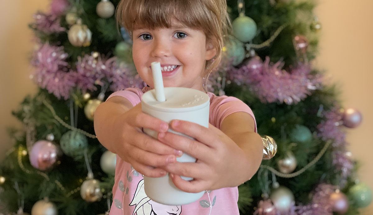 Niña sosteniendo una taza de batido para niños en las manos frente al árbol de Navidad
