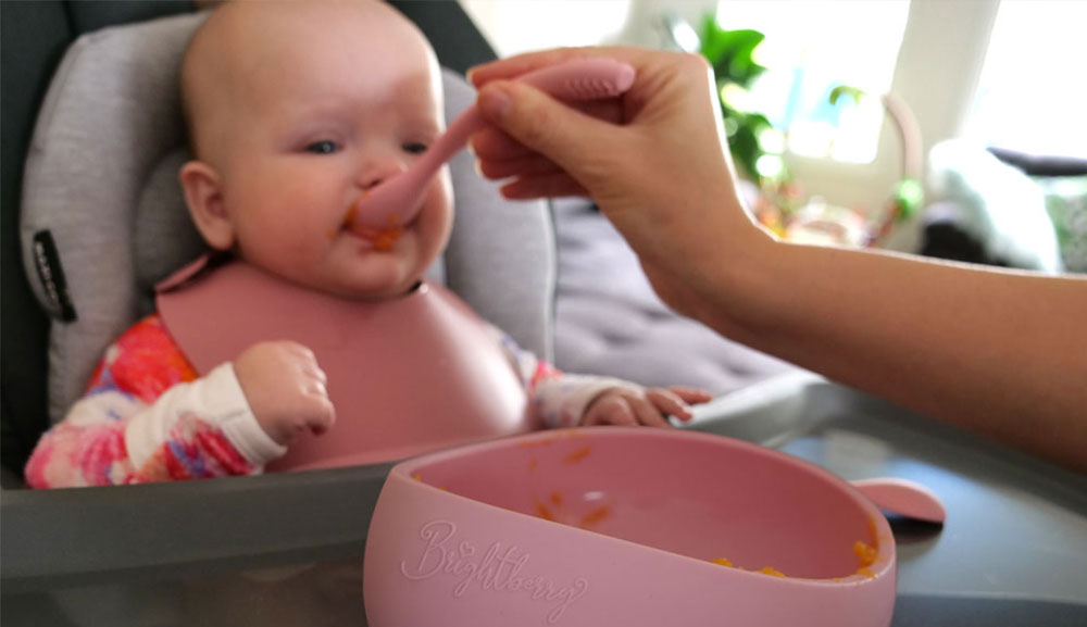 Mamá alimenta a su bebé con puré de calabaza con una cuchara y un tazón Brightberry