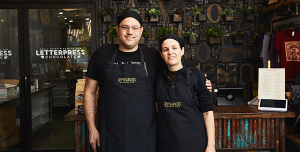A man, David and a woman, Corey stand in the retail area of LetterPress Chocolate, with the text logo to the left on the glass window