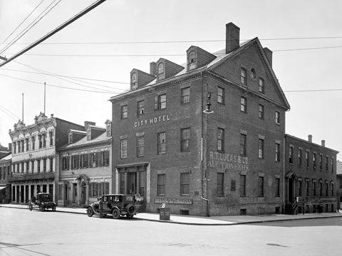 A black and white photograph of a vintage Ford parked in front of Gadsby's in Alexandria