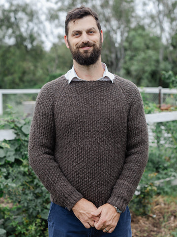 bearded man in his 30s with a slight smile and clasped hands looking at the camera while wearing a handspun and knit sweater. He is standing in front of a garden