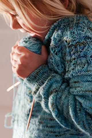 a young blonde girl stands sideways to the camera joyfully holding a ball of yarn attached to the unfinished sweater she is trying on for fit