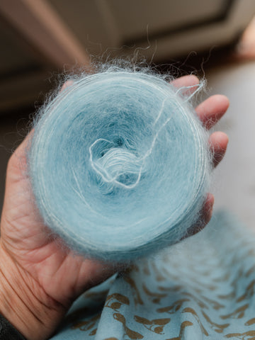 a woman's hand holds a cake of yarn in a soft blue/green colour