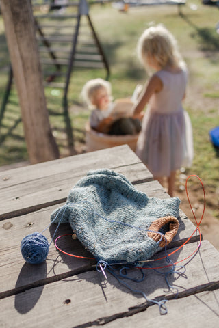 unfinished knitting (a children's vest) sits on a table as children play in the background