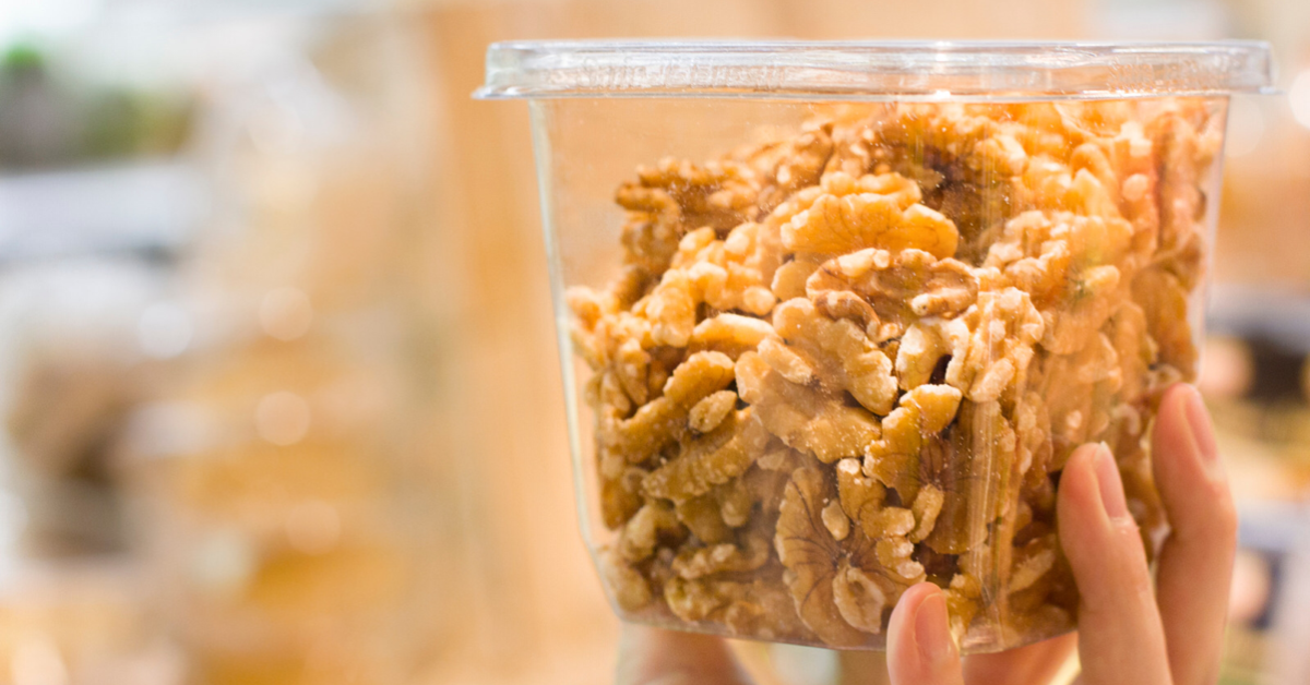 A hand holding up a square see-through container of shelled walnuts against a blurry background