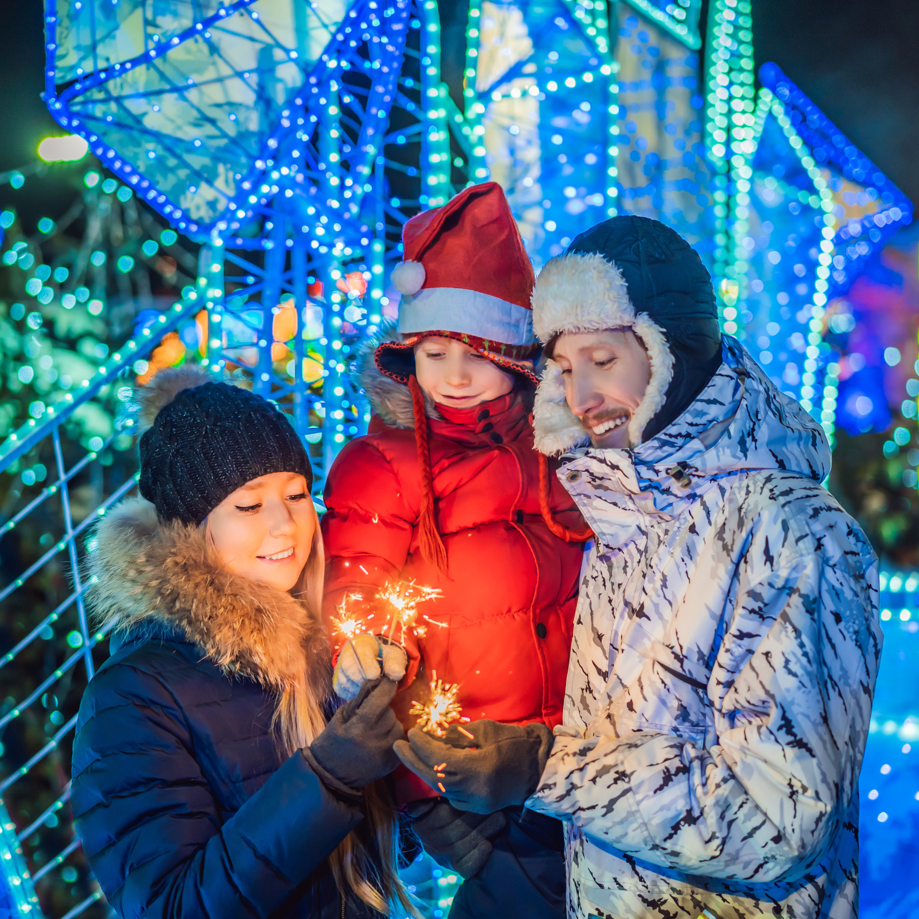 A family of three, warmly dressed in winter coats and hats, stands close together in front of vibrant holiday lights. The child, wearing a Santa hat, holds sparklers while smiling with their parents. The background is illuminated with festive blue and green lights, creating a joyful atmosphere.