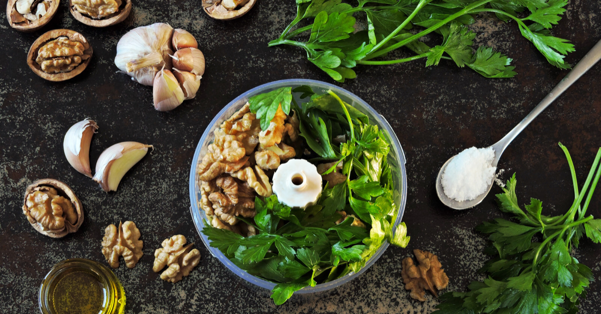 Shelled walnuts and fresh spinach in a food processor on a dark table surrounded by garlic cloves, various herbs and a teaspoon of salt