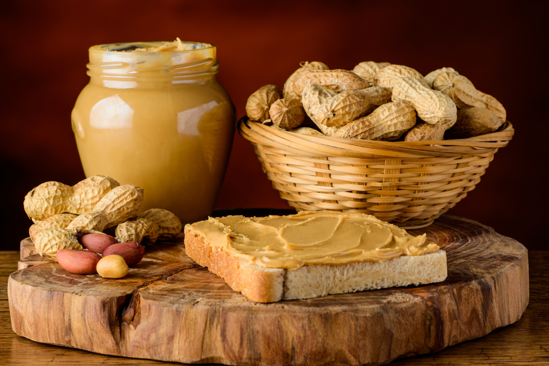 A jar of peanut butter, a basket of unshelled peanuts and a slice of toast with peanut butter on a wooden chopping board.