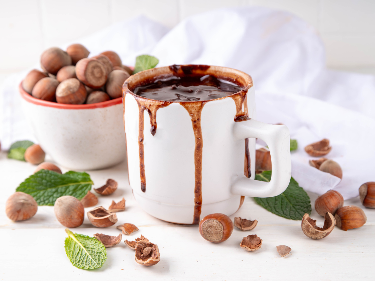 A cup full of delicious homemade hazelnut syrup with a bowl and some loose hazelnuts in the background.