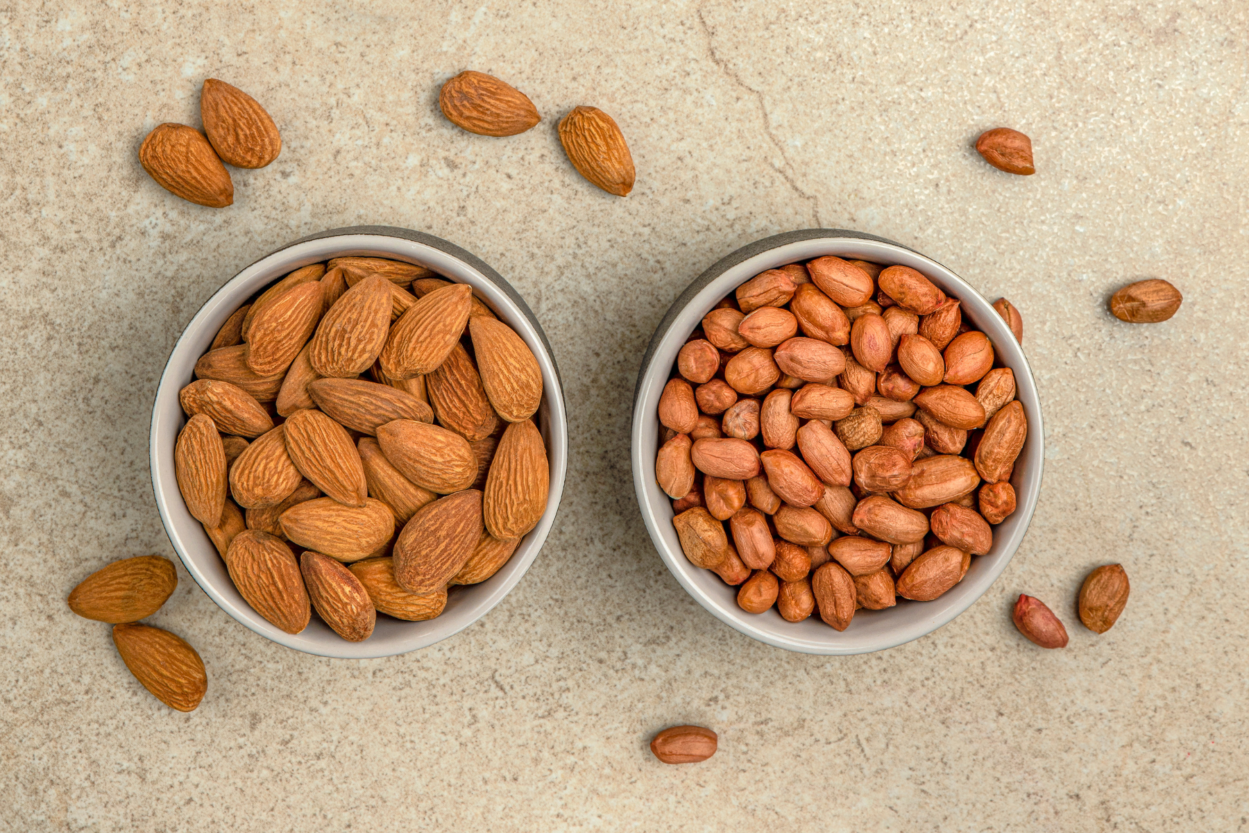 Two bowls on a kitchen table, one filled with raw almonds and one with raw peanuts.