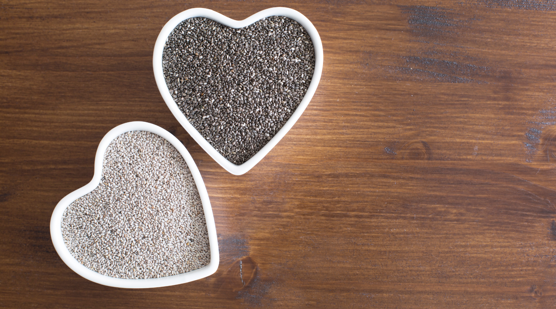Chia seeds in heart-shaped bowls on a wooden table, with white and black chia seeds side by side.