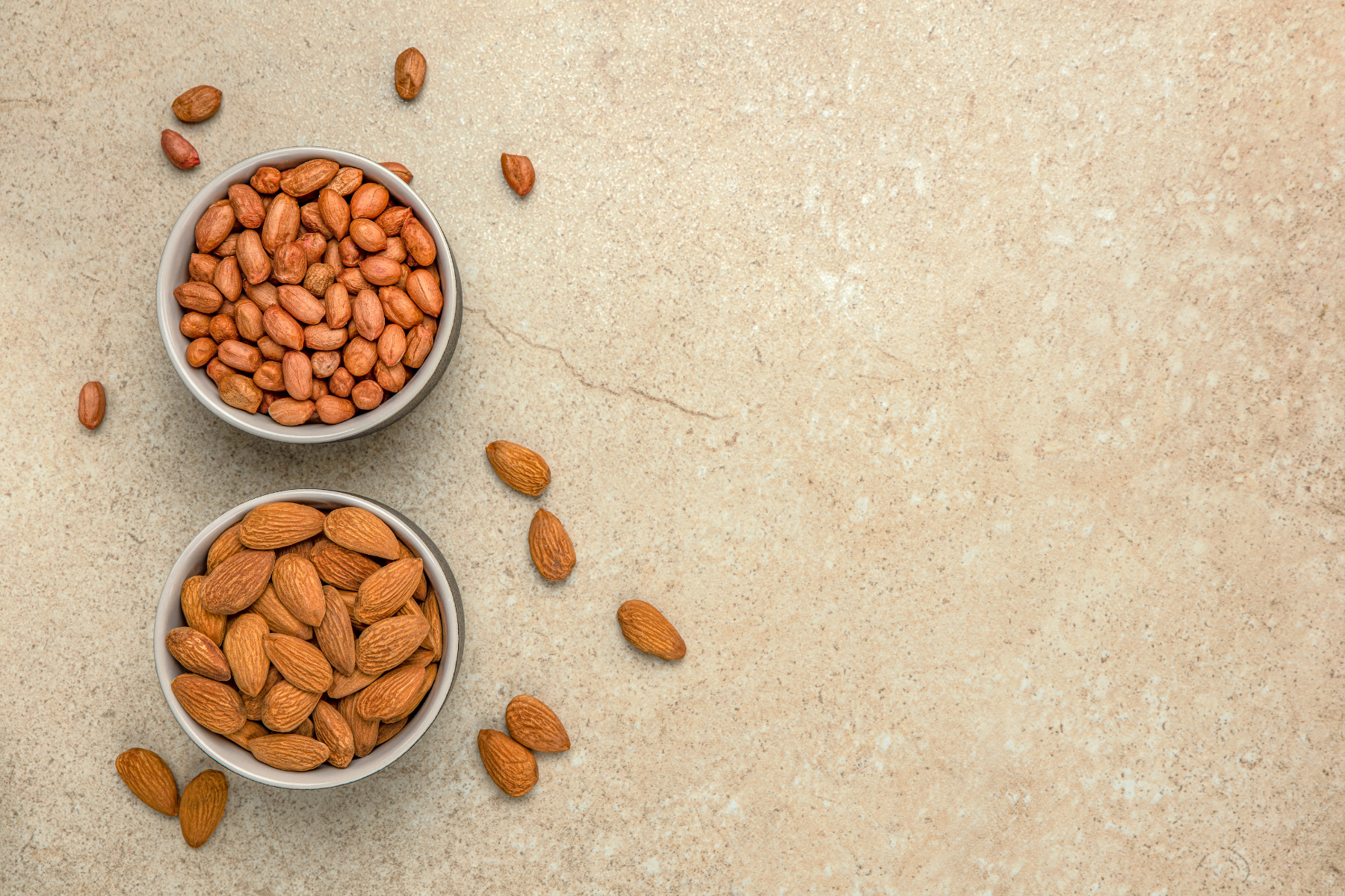 A bowl of almonds next to a bowl of peanuts on a wooden table.