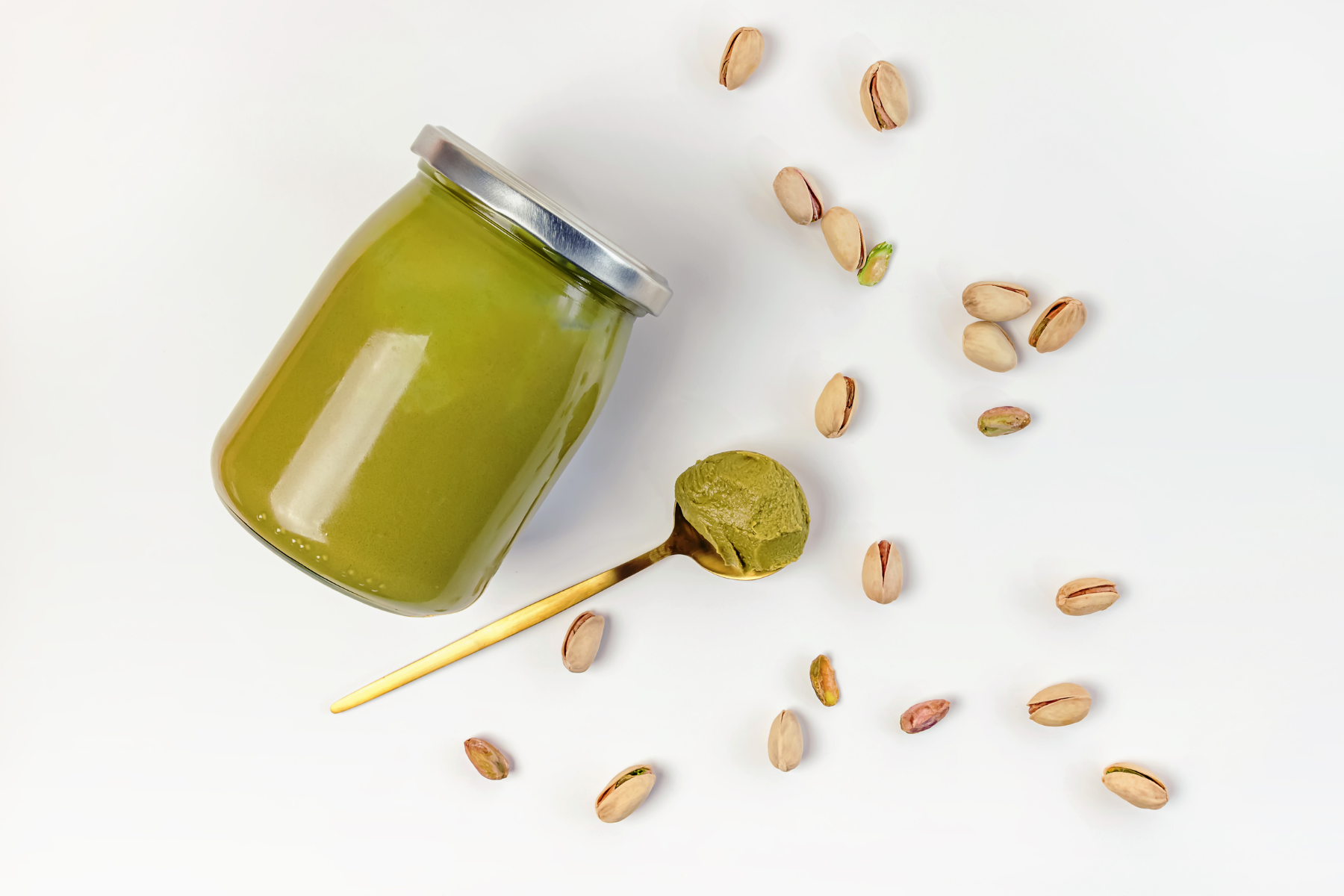A jar of pistachio paste next to some loose pistachio against a white background.