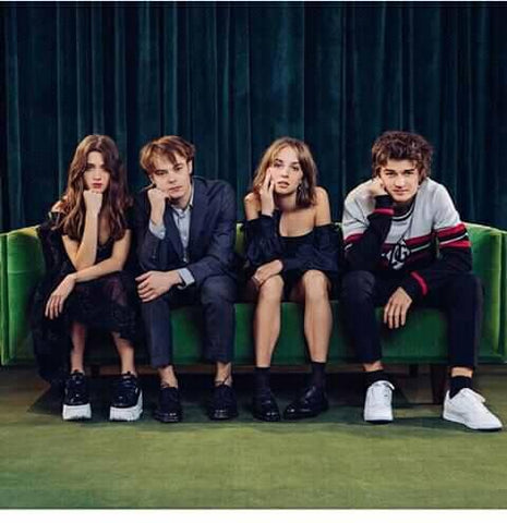 Two white young adult girls and two white young adult boys (sitting girl-boy-girl-boy) sit together on a green couch wearing modern clothing looking directly at the camera in expressions of tiredly waiting for something to arrive.