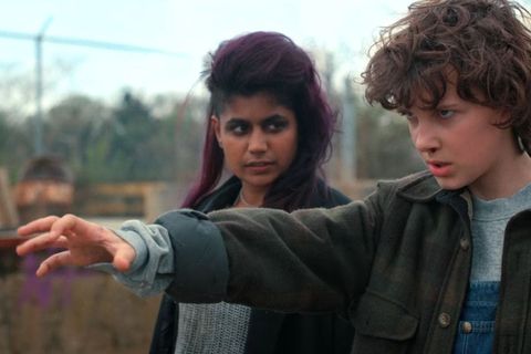 A young brown girl with purple hair and dark eyeliner watches a white girl with short, curly hair standing in a power position with her arm outstretched 