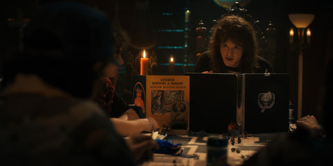 An older white teenage boy with dark, wild hair sits in front of another teenage boy from over a Dungeons and Dragons book in a dark-lit room with the board game set up between them.