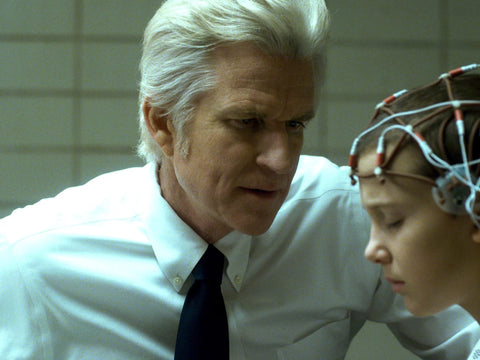 Older white man with silver white hair dressed in a white dress shirt and red tie hovers over young white girl with brain monitors scattered over her shaved head