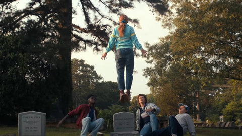 A young white girl wearing jeans and a light blue jacket with colorful stripes in the center is suspended in the air above three teenagers (two white boys and one Black boy) on the ground of a cemetery surrounded by trees. 