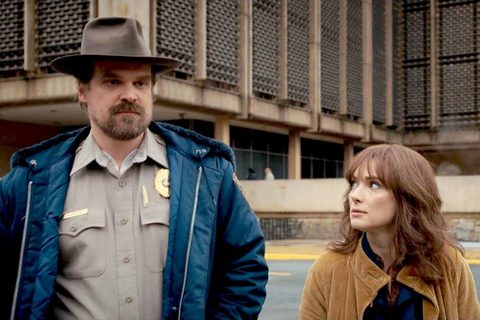 A tall white man with a goatee in his late 50s wearing a dark blue jacket over a ranger uniform walks alongside a considerably shorter white woman with brunette hair cut in the style of bangs wearing a yellow jacket over a dark top