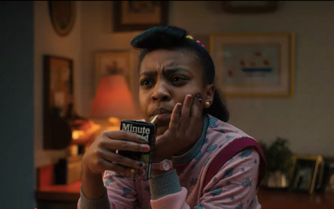 Young teenage Black girl in 70s fashion sips on a juice box while holding her face in one hand, looking suspicious 