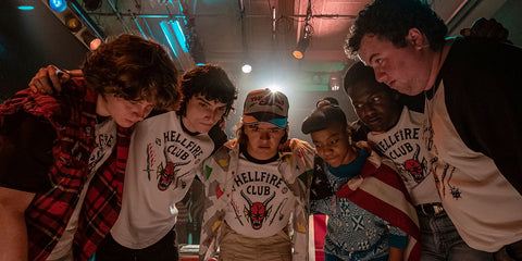 Six teenagers (one Black boy, one Black girl, and four white boys) stand in a circle around a board game wearing matching t shirts that say "Hellfire Club"