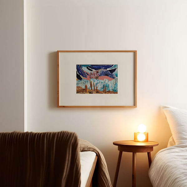 Collage art print by Esme Rose Marsh, featuring torn blue and coral paper, photos of the sea, ice glaciers, cacti in the desert and pink and blue crystals. Art print is in a light wood frame with a large off-white border on a white wall in a room with mid-century style furniture.