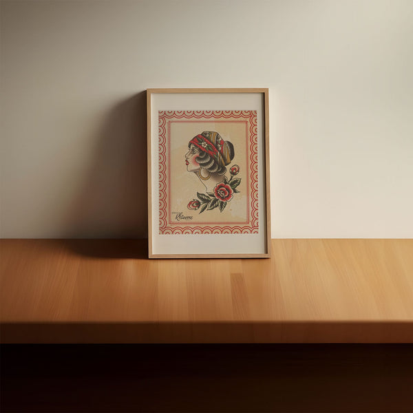 Traditional tattoo style illustration of profile of a woman’s face and neck on a tea stained background. The drawing is very detailed, featuring bold gradients and highlighting red and blue elements throughout. The woman is wearing a head scarf, with large gold hoop earrings and there are red flowers at the base of her neck. in a light wood frame on a wooden table, leaning against a white wall.