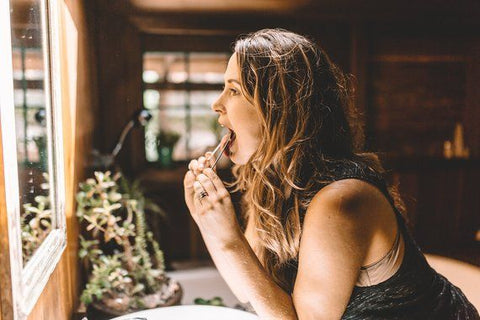 a woman looks into a mirror above a sink whilst she scrapes her tongue with an Ayurvedic copper tongue scraper