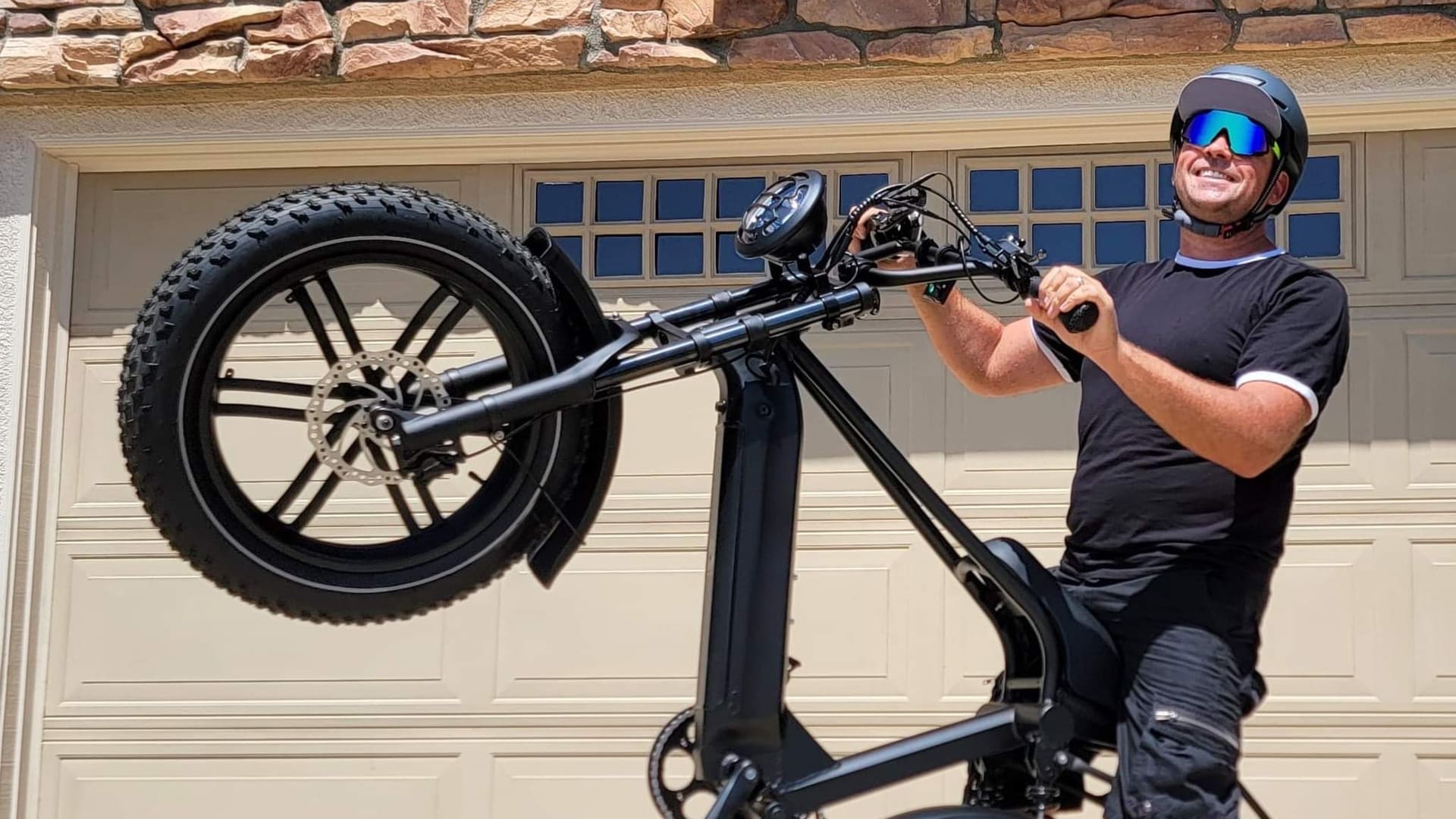 Man wearing an Xnito helmet and sunglasses performing a wheelie on a fat-tire eBike in front of a garage, smiling under the sun