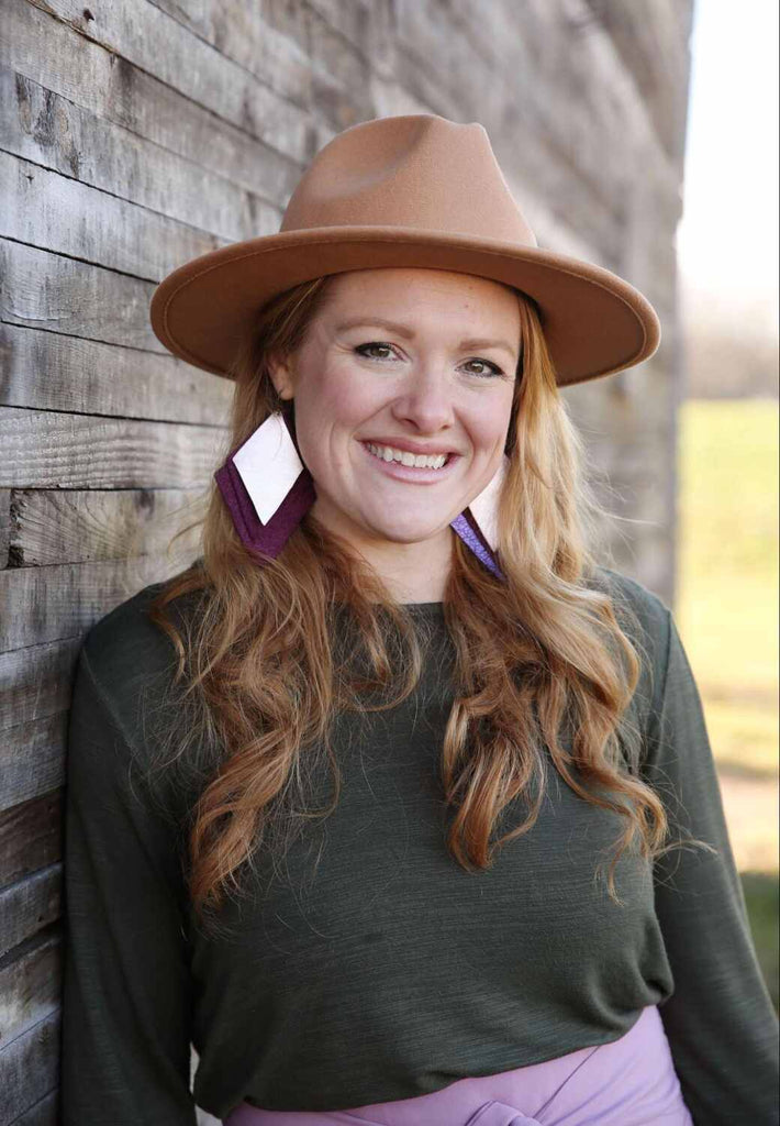 Woman with long red hair stands in front of a wooden paneled wall wearing a great shirt, large earrings, and a tan hat