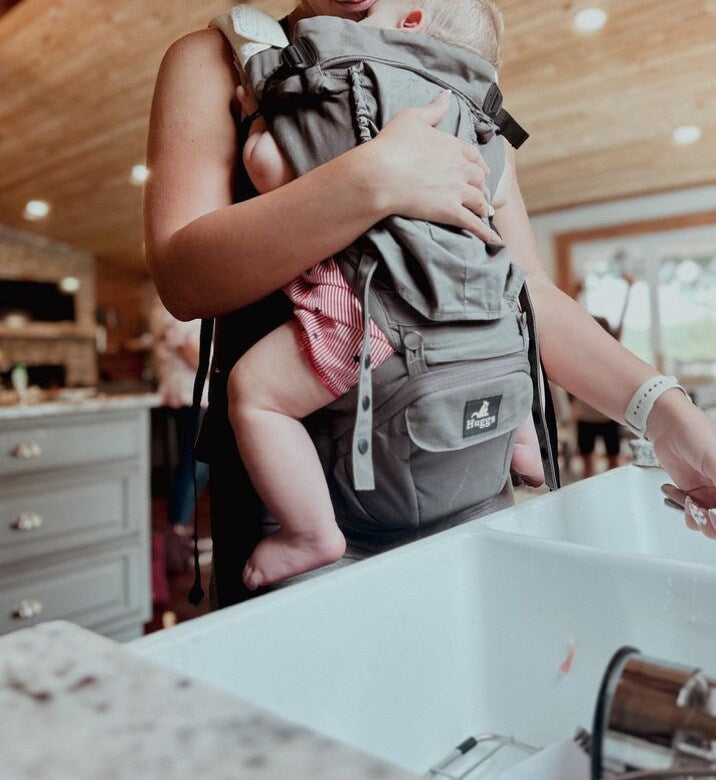 Mom standing in front of a sink holding a baby in the Huggs Carrier