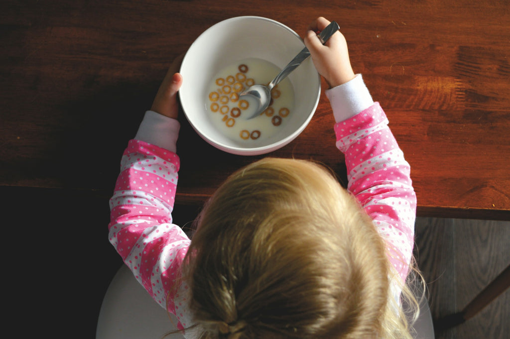 toddler in pink and white shirt sits in front of a bowl of Cheerios on a brown table&nbsp;
