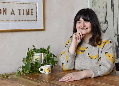 A picture of Kim, sat at a table resting her face on one hand, with the other on the table. There is a fuckoffee cup and a tote vase with a stunning plant in, also on the table. Kim is wearing a jumper with bananas on and is smiling.