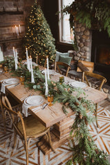 Woodland Christmas Table Setting: a cozy cottage setting with a large wooden dinning table and chairs. There is a Christmas tree and fireplace in the background. The table has minimal decorations with white plates, silver cutlery, white taper candles, orange wine glasses, and a garland of pine and eucalyptus as the table runner.  
