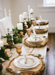 Woodland Christmas Table Setting: wooden table with wooden place mats, white fine china dishes,  copper cutlery, and a garland of fresh greenery as the table runner. The garland has pine cones and twigs intertwined. Mix matched candle holders with tapper and rounded candles. 