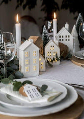 Traditional Christmas Table Setting: white linen table cloth, white plates, and white taper candles. The centrepiece is small, white festive houses with miniature Christmas tree sculptures.