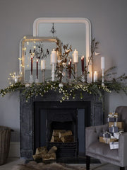 Woodland Christmas mantel decorations: a black mantel with pine branches and babys breath on top,  there is a mix of tall white and glass candle holders with red and white taper candles.  There are wild branches sticking out. There are two mirrors sitting on the mantel with fairy lights draping over one. There is a grey chair beside the mantel with small gifts on it.