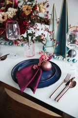 A Merry and Bright Christmas Table Setting: a white table with metallic cutlery, navy blue plates, purple linen napkins, a jewel toned flower centrepiece, and glass teal Christmas tree sculptures. There is a garland of small teal beads. 