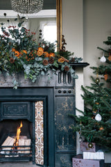 A black, vintage mantel decorated with woodland inspired Christmas decorations. There is a large centrepiece with eucalyptus, pine, dried oranges, berries, and other greenery.  There is a large gold mirror behind the centrepiece. Off to the side is a decorated Christmas tree. 