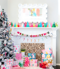 A Merry and Bright Christmas mantel: a frosted Christmas tree to the side with youthful ornaments, several gifts with bright wrapping paper, and a fireplace filled with logs of wood. On top of the white mantel are vibrant and pastel ceramic Christmas trees and a white, modern mirror. A "merry christmas" banner and candy cane garland are hanging from side to side.