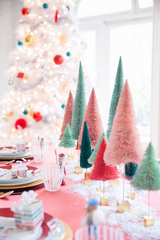 A Merry and Bright Christmas Table Setting: a pink table cloth, fake snow as the table runner, and an assortment of pastel brush bottled trees. There are red and gold plates and a blurred white Christmas tree in the background.